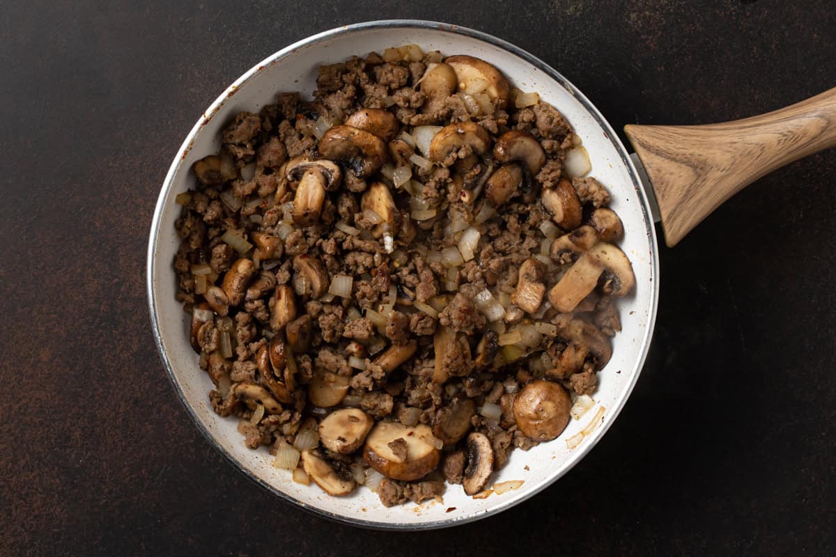 Browned ground sausage, sliced mushrooms and chopped onions in a white skillet.