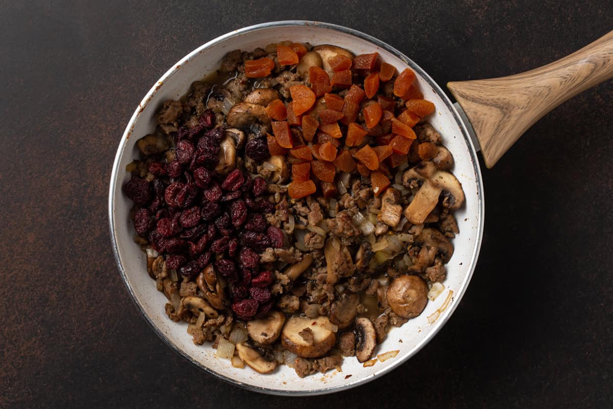 Browned ground sausage, mushrooms, onions, dried cranberries and dried apricots in a white sauté pan.