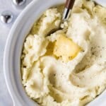 Mashed potatoes with a melting pat of butter, cracked black pepper, and a serving spoon in a white slow cooker.