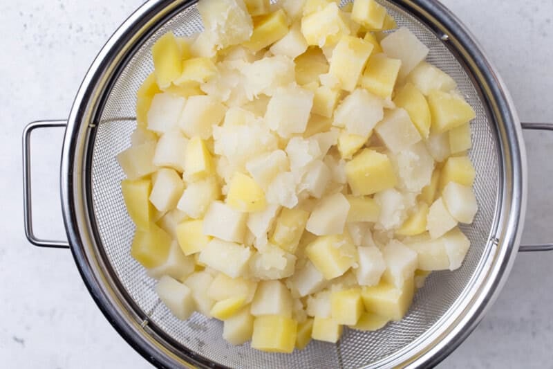 Boiled potato cubes draining in a mesh strainer, tender and just starting to soften at the edges.