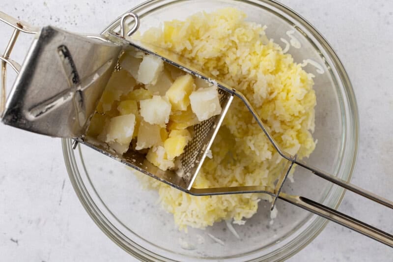 Cooked potato cubes being pressed through a potato ricer into a glass bowl of fluffy riced potatoes.