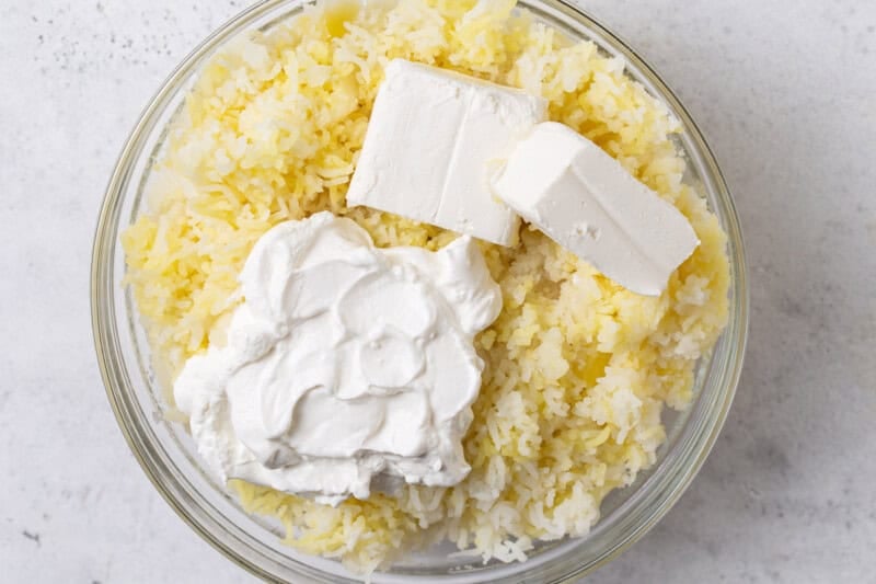Riced potatoes in a glass bowl topped with a dollop of sour cream and two blocks of cream cheese, ready to mix.
