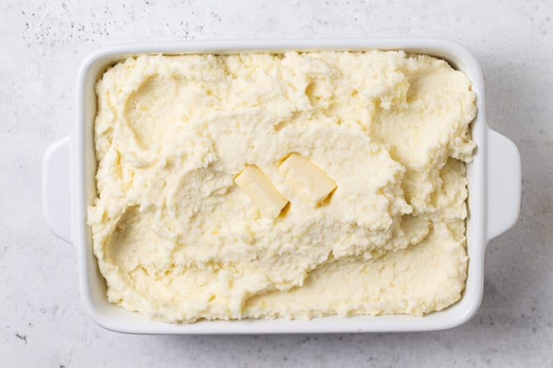 Mashed potatoes spread in a white baking dish with two pats of butter on top, ready to cover and refrigerate.